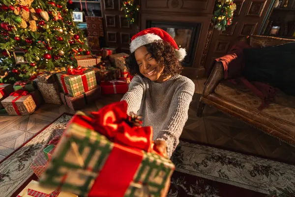 Merry Christmas. African American woman giving you present gift box near Christmas tree. Girl in living room with Christmas tree and fireplace packing wrapping opening gift box. Christmas eve at home