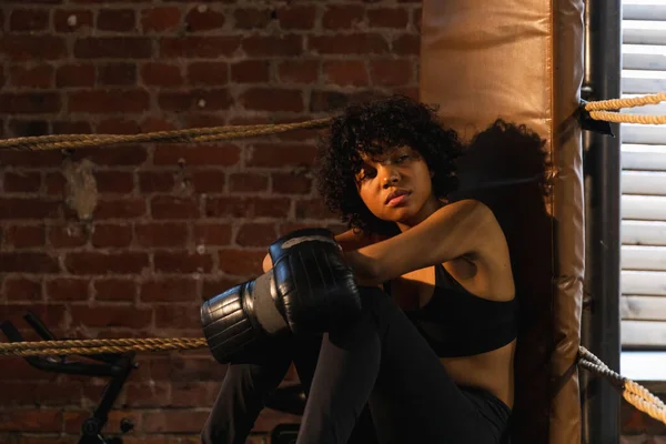 Woman fighter girl power. African american woman fighter with boxing gloves sitting on boxing ring waiting and resting after fight. Strong powerful girl in gym. Strength fit body workout training