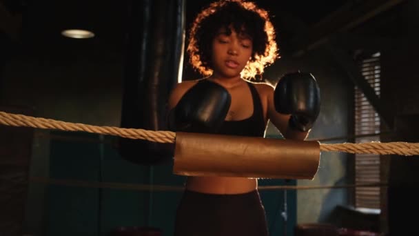 女战士女孩的力量 拿着拳击手套站在拳击台上 靠着绳索等待休息的非裔美国妇女战士 强壮有力的女孩 力量适合体能训练 — 图库视频影像