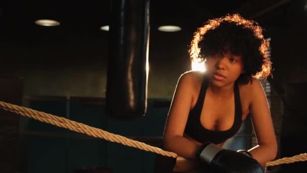 女戦士ガールパワー ロープを待って休憩にもたれてボクシングリングに立っているボクシングの手袋を持つアフリカ系アメリカ人女性の戦闘機 強い力を持つ少女 筋力トレーニング フィットボディトレーニング — ストック動画