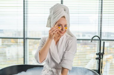 Göz bandı güzellik tedavisi. Göz altları kolajen pedli bir kadın. Kafasında göz bandı olan kız yüz bakımı yaptırıyor. Bornozlu, havluyla banyoda dinlenen genç ve güzel bir kadın.