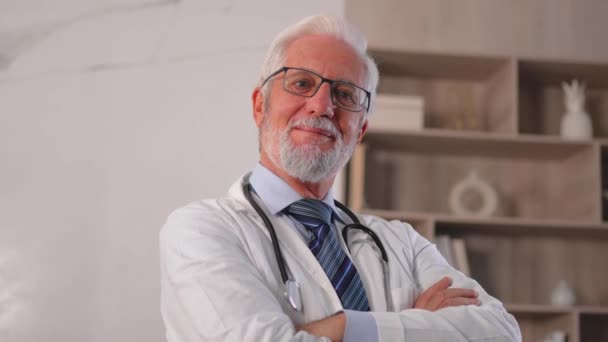 メガネの熟年男性医師 病院や医師のオフィスでカメラを見ている笑顔の医学ユニフォーム Gpセラピスト クリニックの医療専門家 ヘルスケア — ストック動画