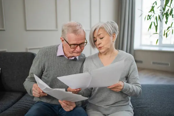 Sad tired disappointed middle aged senior couple sit with paper document. Unhappy older mature man woman reading paper bill managing bank finances calculating taxes planning loan debt pension payment
