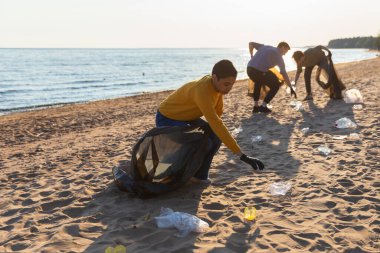 Dünya günü. Gönüllü eylemciler sahil bölgesindeki çöpleri topluyor. Kadınlar ve erkekler okyanus kıyısındaki çöp torbasına plastik çöp koyar. Çevre koruma kıyı bölgesi temizliği