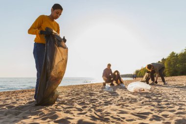 Dünya günü. Gönüllü eylemciler sahil bölgesindeki çöpleri topluyor. Kadınlar ve erkekler okyanus kıyısındaki çöp torbasına plastik çöp koyar. Çevre koruma kıyı bölgesi temizliği