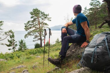 Uçuruma tırmanma macerası. Gezintiden sonra güzel manzaraya bakan sırt çantalı adam dinleniyor. Sırt çantalı gezgin yeşil orman ve gölün üzerindeki büyük kayanın üzerinde oturuyor. Yürüyüşçü adam turizmin tadını çıkarıyor.