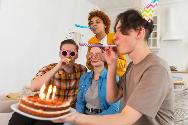 Make a wish. Woman wearing party cap blowing out burning candles on birthday cake. Happy Birthday party. Group of friends wishes girl happy birthday. People celebrating birthday with party at home