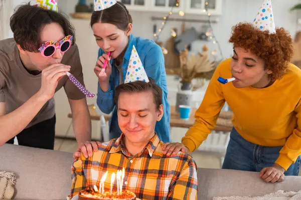 Make a wish. Man wearing party cap blowing out burning candles on birthday cake. Happy Birthday party. Group of friends wishes guy happy birthday. People celebrating birthday with party at home