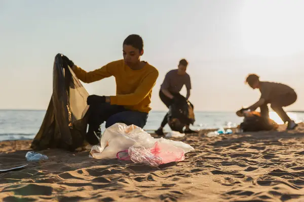 Earth day. Volunteers activists collects garbage cleaning of beach coastal zone. Woman mans with trash in garbage bag on ocean shore. Environmental conservation coastal zone cleaning. Blurred image