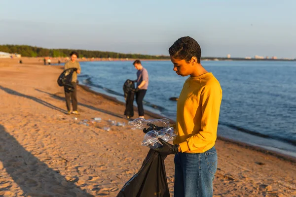 Earth day. Volunteers activists team collects garbage cleaning of beach coastal zone. Woman puts plastic bottle trash in garbage bag on ocean shore. Environmental conservation coastal zone cleaning