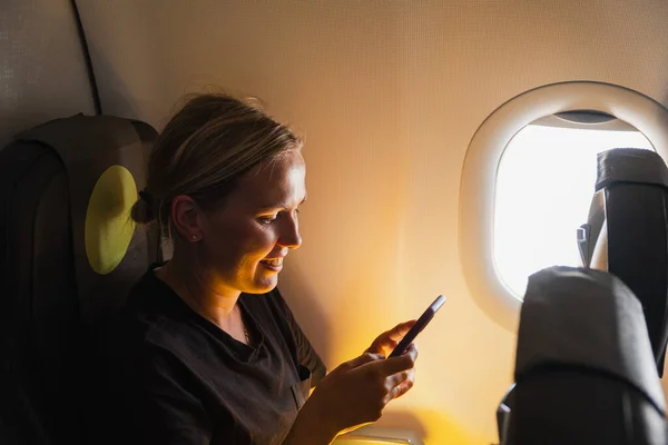 Passenger woman is flying in plane. Girl using phone sitting by airplane window. Traveling female with smartphone inside plane enjoying flight. Woman using wifi connection on board. Traveling girl