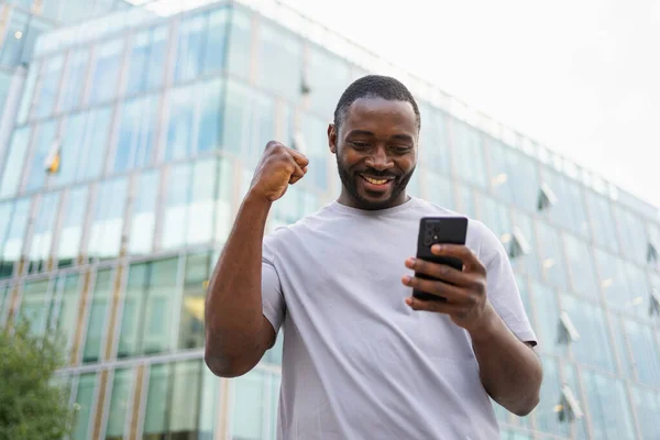Happy African American man euphoric winner with smartphone on street in city. Person guy looking at cell phone reading great news getting good result winning online bid feeling amazed. Winning gesture