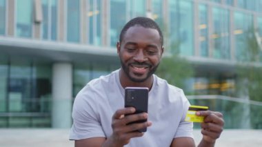 Çevrimiçi alışveriş yapan Afro-Amerikalı adam elinde altın kredi kartıyla alışveriş yapıyor. Şehirdeki sokaktaki adam internet üzerinden alıyor. Kredi kartı detaylarını gir. Online alışveriş ekommerce dağıtım servisi.