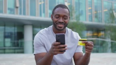 Çevrimiçi alışveriş yapan Afro-Amerikalı adam elinde altın kredi kartıyla alışveriş yapıyor. Şehirdeki sokaktaki adam internet üzerinden alıyor. Kredi kartı detaylarını gir. Online alışveriş ekommerce dağıtım servisi.