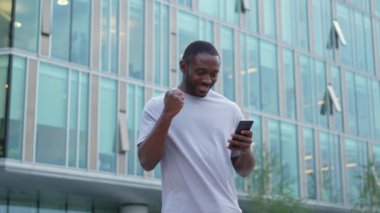 Şehirde akıllı telefonlu mutlu Afrikalı Amerikalı adam mutluluktan havalara uçuyor. Cep telefonuna bakan adam harika haberler okuyor ve online ihaleyi kazanırken çok şaşırıyor. Kazanan jest