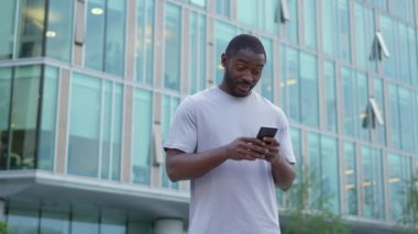 Şehirde akıllı telefonlu mutlu Afrikalı Amerikalı adam mutluluktan havalara uçuyor. Cep telefonuna bakan adam harika haberler okuyor ve online ihaleyi kazanırken çok şaşırıyor. Kazanan jest