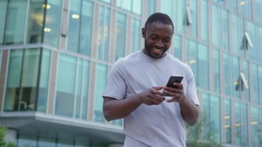 Şehirde akıllı telefonlu mutlu Afrikalı Amerikalı adam mutluluktan havalara uçuyor. Cep telefonuna bakan adam harika haberler okuyor ve online ihaleyi kazanırken çok şaşırıyor. Kazanan jest