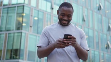 Şehirde akıllı telefonlu mutlu Afrikalı Amerikalı adam mutluluktan havalara uçuyor. Cep telefonuna bakan adam harika haberler okuyor ve online ihaleyi kazanırken çok şaşırıyor. Kazanan jest