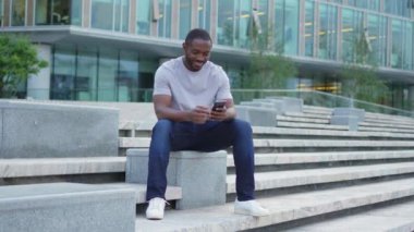Şehirde akıllı telefonlu mutlu Afrikalı Amerikalı adam mutluluktan havalara uçuyor. Cep telefonuna bakan adam harika haberler okuyor ve online ihaleyi kazanırken çok şaşırıyor. Kazanan jest