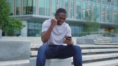 Şehirde akıllı telefonlu mutlu Afrikalı Amerikalı adam mutluluktan havalara uçuyor. Cep telefonuna bakan adam harika haberler okuyor ve online ihaleyi kazanırken çok şaşırıyor. Kazanan jest