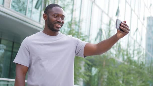 スマートフォンを持っているアフリカ系アメリカ人男性は 都市の街頭でビデオチャットを持っています ソーシャルネットワークで最高の友人とチャットガイブロガー バーチャル会議オンラインチャットビデオコール屋外を持っている男 — ストック動画
