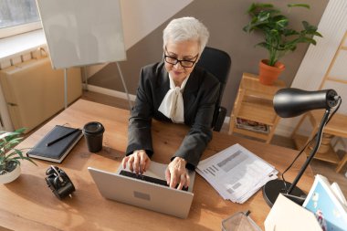 Confident stylish european middle aged senior woman using laptop at workplace. Stylish older mature 60s gray haired lady businesswoman sitting at office table. Boss leader teacher professional worker