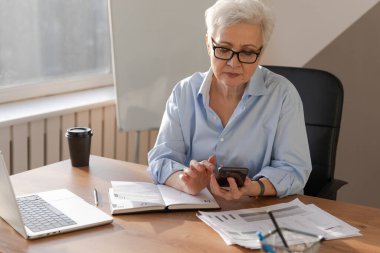 Confident stylish european middle aged senior woman using smartphone at workplace. Stylish older mature 60s gray haired lady businesswoman with cell phone in office. Boss leader using internet apps