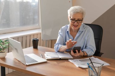 Confident stylish european middle aged senior woman using smartphone at workplace. Stylish older mature 60s gray haired lady businesswoman with cell phone in office. Boss leader using internet apps