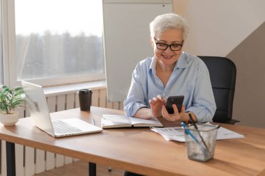 İş yerinde akıllı telefon kullanan kendine güvenen Avrupalı orta yaşlı bir kadın. Tarz sahibi, olgun, 60 'lı yaşlarda, ofisinde cep telefonu olan beyaz saçlı bir iş kadını. Patron internet uygulamaları kullanıyor