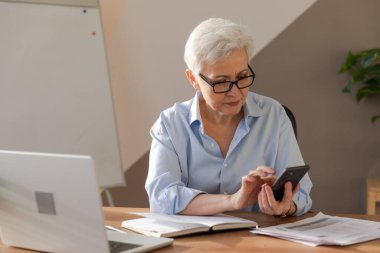 Confident stylish european middle aged senior woman using smartphone at workplace. Stylish older mature 60s gray haired lady businesswoman with cell phone in office. Boss leader using internet apps