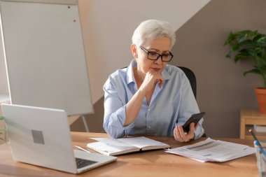 Confident stylish european middle aged senior woman using smartphone at workplace. Stylish older mature 60s gray haired lady businesswoman with cell phone in office. Boss leader using internet apps