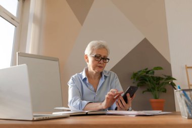 Confident stylish european middle aged senior woman using smartphone at workplace. Stylish older mature 60s gray haired lady businesswoman with cell phone in office. Boss leader using internet apps