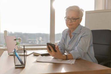 Confident stylish european middle aged senior woman using smartphone at workplace. Stylish older mature 60s gray haired lady businesswoman with cell phone in office. Boss leader using internet apps