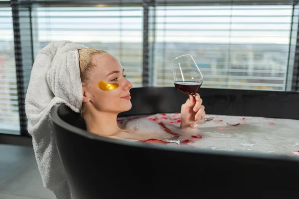 Spa relaxation. Woman lying in bath with holding glass with red wine. Girl relaxing in bathroom at home. Pretty female taking hot bath drinking wine. Stress relief. Rest after hard working day