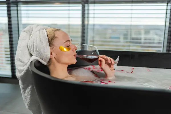 Spa relaxation. Woman lying in bath with holding glass with red wine. Girl relaxing in bathroom at home. Pretty female taking hot bath drinking wine. Stress relief. Rest after hard working day