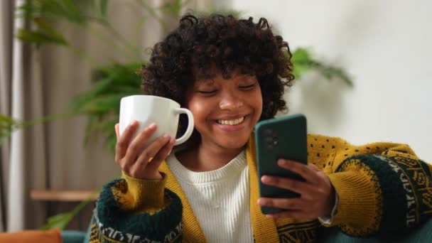 早上好非洲女孩拿着智能手机喝咖啡 坐在家里的沙发上 女人用手机上网 使用社交媒体应用程序 网上购物网上新闻手机成瘾 — 图库视频影像