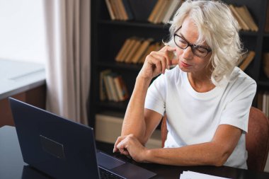 İş yerinde dizüstü bilgisayar kullanan kendine güvenen orta yaşlı bir kadın. Yaşlı, olgun bir iş kadını, ofis masasında dizüstü bilgisayar ve kağıt belgelerle oturuyor. Patron öğretmen profesyonel işçi.