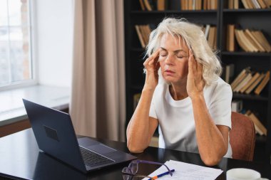 Baş ağrısı. Orta yaşlı bir kadın tapınaklara dokunuyor ve stres yaşıyor. Yaşlı kadın baş ağrısı hissetmekten yoruldu şakakların alnına sürtünmekten. Uzun dizüstü bilgisayar kullanımı ofis vizyonu sendromu