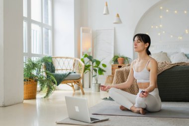 Yoga farkındalığı meditasyonu çevrimiçi. Evde dizüstü bilgisayar dersleriyle yoga yapan bir kadın. Lotus 'ta oturan kadın içeride meditasyon yapıyor. Nefes egzersizi yapan bir kız. Evde yoga.