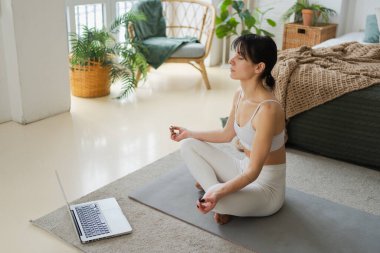 Yoga farkındalığı meditasyonu çevrimiçi. Evde dizüstü bilgisayar dersleriyle yoga yapan bir kadın. Lotus 'ta oturan kadın içeride meditasyon yapıyor. Nefes egzersizi yapan bir kız. Evde yoga.