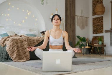 Yoga farkındalığı meditasyonu çevrimiçi. Evde dizüstü bilgisayar dersleriyle yoga yapan bir kadın. Lotus 'ta oturan kadın içeride meditasyon yapıyor. Nefes egzersizi yapan bir kız. Evde yoga.