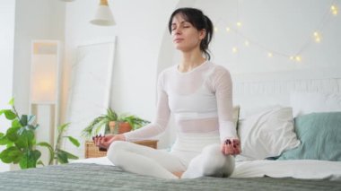Yoga farkındalığı meditasyonu. Sağlıklı bir kadın yatak odasında yoga yapıyor. Nilüfer çiçekli bir kadın yatakta oturmuş, meditasyon yapıyor, gülümsüyor, evde dinleniyor. Nefes egzersizi yapan bir kız. Evde yoga.
