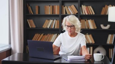 İş yerinde dizüstü bilgisayar kullanan kendine güvenen orta yaşlı bir kadın. Yaşlı, olgun bir iş kadını, ofis masasında dizüstü bilgisayar ve kağıt belgelerle oturuyor. Patron öğretmen profesyonel işçi.