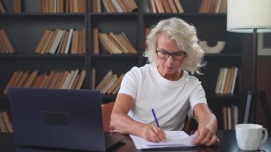İş yerinde dizüstü bilgisayar kullanarak not yazan orta yaşlı bir kadın. Yetişkin, yaşlı bir iş kadınının ofiste çalışıp, online işler yaparak yazılı plaklar çıkarmasına odaklanmış. Olgun patron profesyonel işçi.