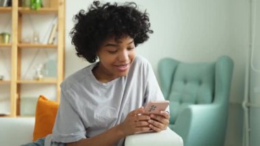 African girl holding smartphone touch screen typing scroll page at home. Woman with cell phone surfing internet using social media apps playing game. Shopping online Internet news cellphone addiction