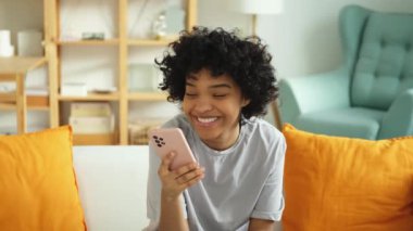 Cep telefonuyla gülen heyecanlı, mutlu, genç siyahi, Afro-Amerikalı kadın. Cep telefonu mesajı almaktan zevk alıyor. Neşeli kız yüksek sesle gülüyor. Kanepede oturup komik video haberleri izliyor.