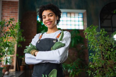 Kendi işimi yürütüyorum. Botanik mağazasında yeşil bitkilerle önlük giyen Afrikalı bir çiçekçi kadın. Çiçekçide çalışan mutlu küçük işletme sahibi etrafı bitkilerle çevrili gülümsüyor.