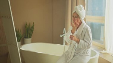 Yetişkin yaşlı bir kadın günlük güzellik rutini yaparak anti-yaş yüz kremi sürüyor. Beyaz maskeli ya da cildinde krem olan yaşlı bir kadın cilt bakımına gidiyor.