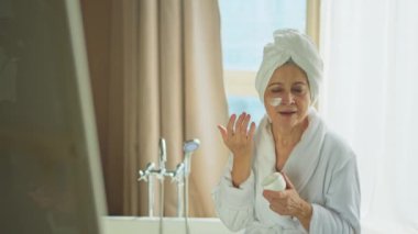 Yetişkin yaşlı bir kadın günlük güzellik rutini yaparak anti-yaş yüz kremi sürüyor. Beyaz maskeli ya da cildinde krem olan yaşlı bir kadın cilt bakımına gidiyor.