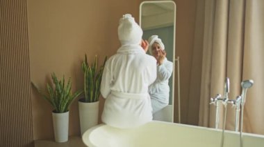 Yetişkin, yaşlı bir kadın aynaya bakıyor ve günlük güzellik kremi sürüyor. Cildinde beyaz maske kremi olan yaşlı bir kadın cilt bakımı görüyor.